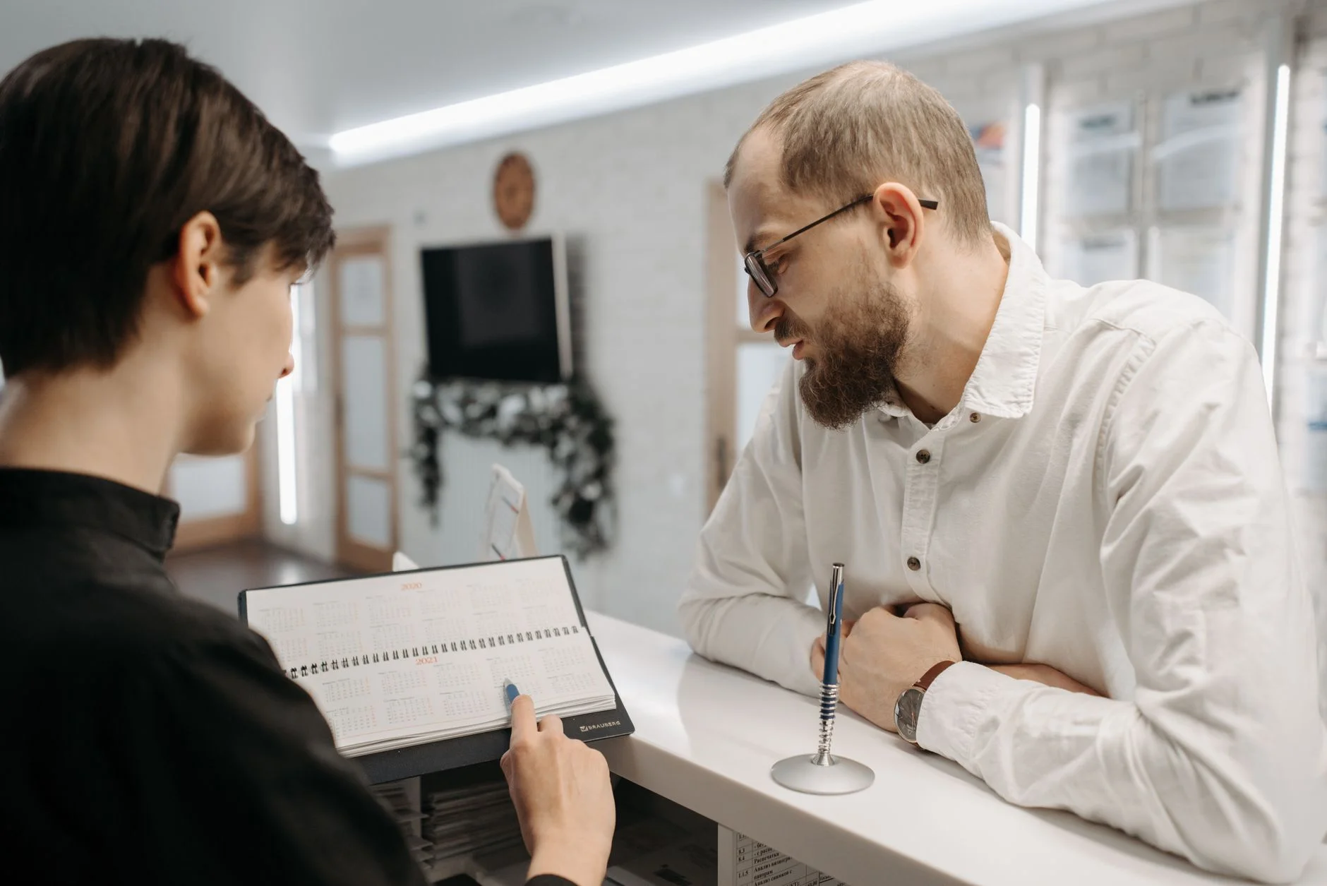 A receptionist and client converse over an appointment book at a clinic reception desk. – Foto: Pavel Danilyuk / Pexels